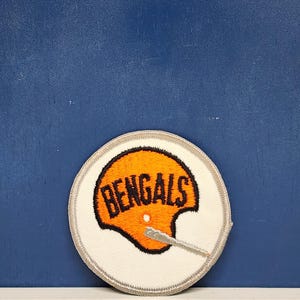 May include: A circular patch featuring the word "BENGALS" in orange lettering on a football helmet design. The patch has a white border with a gray stitched edge. The background is a solid blue.