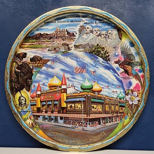 May include: A round metal tray with a colorful design featuring images of Mount Rushmore, the Badlands National Monument, a bison, a pheasant, a flower, and the Corn Palace in Mitchell, South Dakota.