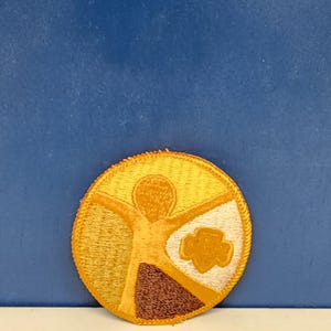 May include: Embroidered patch with a circular design featuring a stylized figure with outstretched arms in shades of yellow and brown. A trefoil emblem is visible on the right side. The patch has a gold border.
