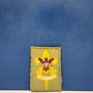 May include: A rectangular khaki-colored embroidered patch featuring a yellow fleur-de-lis emblem with a brown eagle at the center. The eagle holds a shield with red and white stripes. Below the emblem is a yellow banner with the text BE PREPARED.