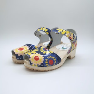 May include: A pair of open-toe clogs with a navy blue base featuring a yellow and pink floral pattern. The shoes have a light-coloured wooden sole, an adjustable ankle strap with a metal buckle, and a white interior label with Swedish flag.