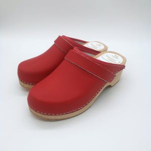 May include: A pair of red leather clogs featuring a wide strap across the top, light-coloured wooden soles, and white stitching along the base. The interior insoles are white with the text Moheda Toffeln and Real Leather printed in black ink.
