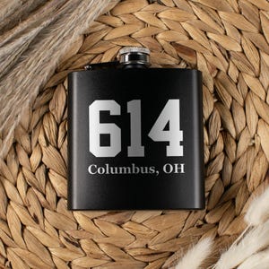 May include: Black flask with a silver cap, featuring the number "614" and the text "Columbus, OH" in white. The flask is set against a woven background with dried plants.