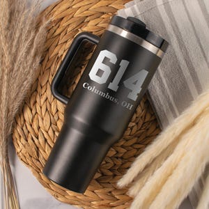 May include: Black travel mug with a handle and a silver rim. The mug features the number "614" in large, silver lettering, with "Columbus, OH" printed below. The mug is set on a woven mat with dried plants and a striped cloth.