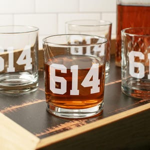 May include: Clear glass tumblers with the number "614" in white. Some glasses are filled with a brown liquid. The glasses are arranged on a dark wooden tray. A bottle with a brown liquid is in the background.