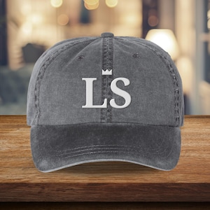 May include: A gray baseball cap with the white letters "LS" and a small crown above the "L". The cap has a curved brim and a slightly faded appearance, suggesting a vintage or worn style. The background is blurred, with warm tones.