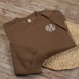 May include: A brown crewneck sweatshirt with a white monogram on the chest. The sweatshirt is folded on a woven mat, with the sleeves and body visible. The monogram features the letters RHG in a circular design.
