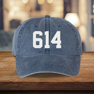 May include: A denim blue baseball cap with the number "614" in bold white lettering across the front. The cap has a curved brim and a fabric strap closure. The hat is sitting on a wooden surface.