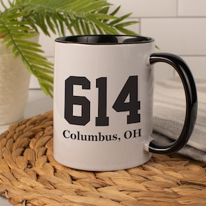May include: White ceramic mug with a black interior and handle. The mug features the number "614" in bold black font, with "Columbus, OH" printed below. The mug is set on a woven mat.
