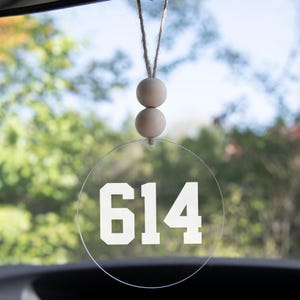 May include: Clear, circular car charm with the white number "614" printed on it. The charm is suspended by a natural-colored string and two wooden beads. The background is a blurred view of green foliage.