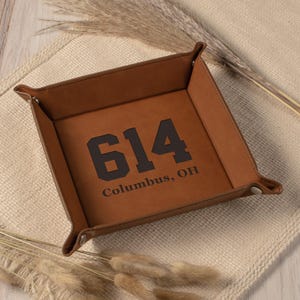 May include: A square, brown leather tray with the number "614" and the text "Columbus, OH" printed in black. The tray has folded corners and is resting on a beige surface.
