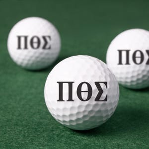May include: Three white golf balls with the Greek letters ΠΘΣ printed in black. The golf balls are on a green surface, with the focus on the front ball. The balls have a dimpled surface.