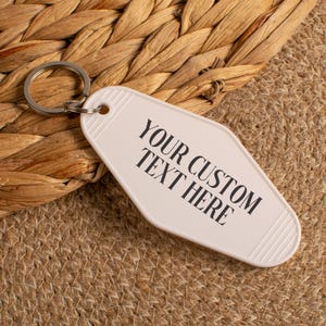 May include: A white, diamond-shaped motel-style keychain with a silver metal split ring. The keychain features the black printed text YOUR CUSTOM TEXT HERE in a serif font, with grooved detailing at the top and bottom edges of the plastic tag.