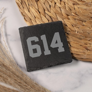 May include: A square, black slate coaster with the number "614" in a light gray, bold font. The coaster is set against a woven background with dried grass.