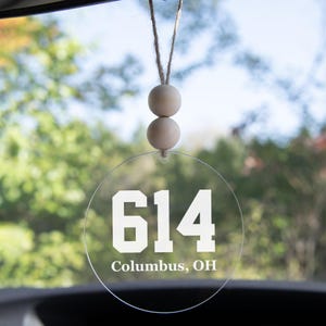 May include: A clear, circular car air freshener with the number "614" and "Columbus, OH" printed in white. It hangs from a string with two wooden beads. The background is a blurred view of green foliage.