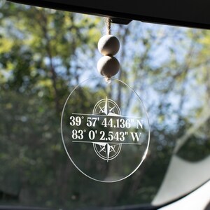 May include: Clear, circular car charm with white text and a compass design. The text reads "39° 57' 44.136" N 83° 0' 2.543" W". The charm hangs from a string with two wooden beads.