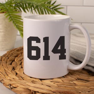 May include: White ceramic mug with a black number 614 printed on the front. The mug has a curved handle and sits on a woven mat. A plant and a gray cloth are in the background.