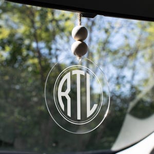 May include: Clear, circular car charm with the initials "RTL" in a white, circular design. The charm hangs from a string with two wooden beads. The background shows a blurred view of trees and sky.
