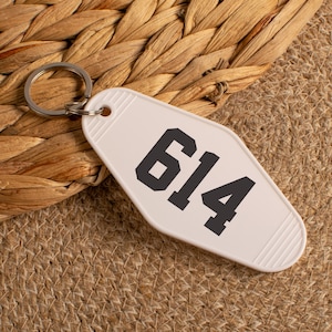 May include: A white motel-style keychain with the number "614" in bold, black font. The keychain has a silver metal ring and is set against a textured, neutral background.