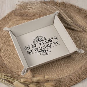 May include: A square, off-white leather tray with a compass design and coordinates printed in black. The tray has rounded corners and is set on a textured, neutral-toned surface. The text on the tray reads: 39° 57' 44.136" N 83° 0' 2.543" W.