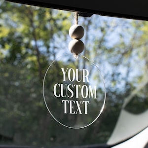 May include: A clear, circular car charm with the text "YOUR CUSTOM TEXT" in white. The charm hangs from a string with two wooden beads. The background shows a blurred view of trees and sky.
