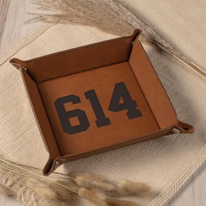 May include: A square, brown leather tray with the number "614" printed in black. The tray has folded corners and is designed to hold small items. The background is a neutral beige.