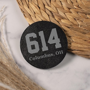 May include: A round, black coaster with the number "614" in gray, along with "Columbus, OH" in a matching font. The coaster is made of a dark, textured material and is set against a woven background.