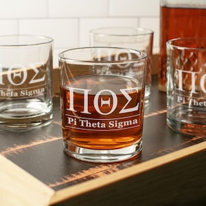 May include: Clear glass tumblers with white Greek letters and the words "Pi Theta Sigma" are filled with amber liquid. The glasses are arranged on a dark wooden tray, with a bottle of amber liquid in the background. The glasses are for drinking.