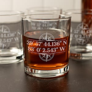 May include: Clear glass tumbler with a compass rose design and coordinates etched in white. The glass is filled with a brown liquid. Other glasses with similar designs are in the background.