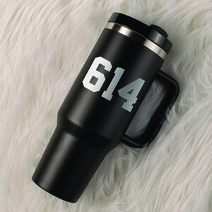 May include: A black travel mug with a handle and a silver rim. The mug features the number "614" in white block letters. The mug is sitting on a white, fluffy surface.