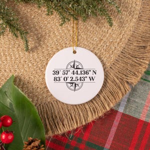 May include: White ceramic Christmas ornament with a compass design and coordinates printed in black. The ornament hangs from a gold string. The background includes a burlap-like material, greenery, and a red plaid pattern.
