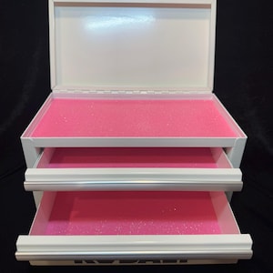 May include: White metal jewelry box with two pull-out drawers and a hinged lid. The interior of the box and drawers are lined with bright pink foam. The box has silver-colored trim.