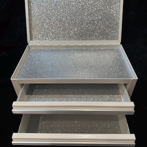 May include: A white, three-tiered storage box with a hinged lid. The interior of the box and drawers are lined with silver glitter. The box is designed to hold small items and is set against a black background.