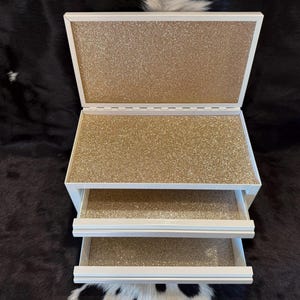May include: A white, three-drawer jewelry box with a hinged lid. The interior and drawer bottoms are covered in gold glitter. The box is open, revealing the glittery interior. The box is sitting on a black and white cowhide.