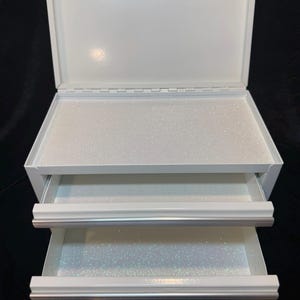May include: White metal storage box with two pull-out drawers and a hinged lid. The interior of the box and drawers have a glittery, iridescent finish. The box has a clean, modern design.