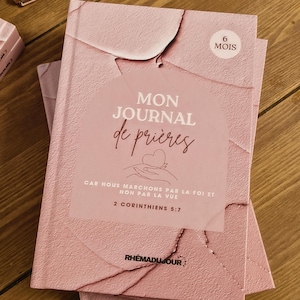 Peut inclure: Deux journaux roses empilés, avec l'inscription "MON JOURNAL de prières" et "6 MOIS" sur la couverture. La couverture comprend également le texte "CAR NOUS MARCHONS PAR LA FOI ET NON PAR LA VUE 2 CORINTHIENS 5:7" et la marque "RHEMADUJOUR".