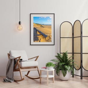 May include: A rocking chair with a grey throw blanket sits in a living room with a framed photograph of a beach scene, a potted plant, and a small side table.