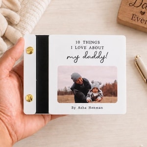 May include: A small, rectangular photo book with a white cover featuring the text 10 THINGS I LOVE ABOUT my daddy! and By Asha Henman. The cover includes a photograph of an adult and a baby, held together by two gold-colored metal binding screws.