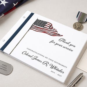 May include: A white rectangular retirement book featuring a graphic of an American flag and the text: Thank you for your service, CONGRATULATIONS ON YOUR RETIREMENT, Colonel James R. Whitaker, and 2015 - 2027, with a dark blue spine.