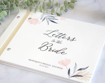 May include: A white rectangular keepsake book with a metal post binding. The cover features a floral design with pink flowers and grey leaves, and the text "Letters to the Bride" followed by "MONIQUE'S BRIDAL SHOWER 02.22.2025" in black script.