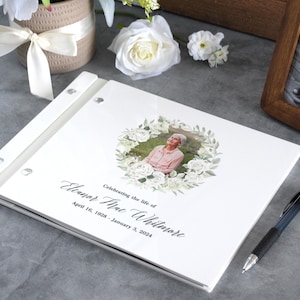 May include: A white memorial guest book featuring a central circular floral wreath surrounding a photograph of a person. The text reads Celebrating the life of Eleanor Mae Whitmore, April 16, 1928 - January 3, 2024. The book has a white spine with silver metal binding screws.