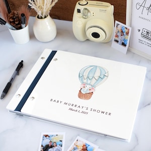 Puede incluir: Un álbum de fotos blanco con lomo azul oscuro y tornillos de encuadernación plateados. La portada presenta una ilustración en acuarela de un elefante en un globo aerostático azul claro y blanco, con el texto BABY MURRAY'S SHOWER y March 1, 2025.