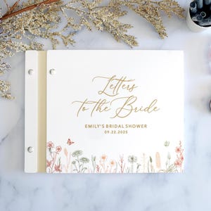 May include: A white rectangular guest book with a gold metal binding spine. The cover features the gold script text Letters to the Bride, Emily's Bridal Shower, and 09.22.2025. A colorful botanical illustration of wildflowers and a butterfly is printed along the bottom edge.