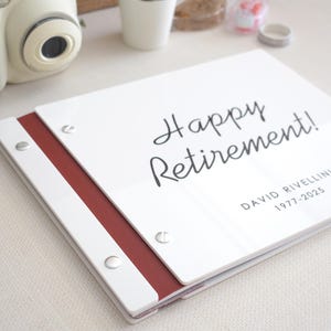 May include: A white rectangular guest book with a dark red spine and silver metal binding screws. The cover features the black printed text Happy Retirement! followed by the name David Rivellini and the years 1977-2025 in a clean font.