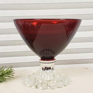 Peut inclure: Un saladier en verre rouge rubis de forme conique évasée. Le saladier repose sur un socle décoratif transparent avec des détails rouges et blancs. La base présente des accents ronds en verre transparent. Le fond est à motifs rayés.