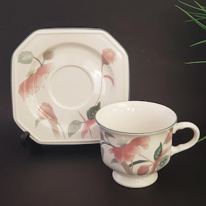 May include: A white teacup and saucer set with a floral design. The cup has a pedestal base and a handle. The saucer is square with rounded corners. Both pieces feature pink flowers and green leaves with a blue trim.