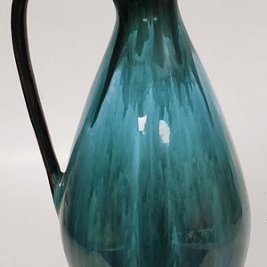 May include: A tall, teardrop-shaped ceramic pitcher with a dark brown rim and handle. The body transitions from a deep teal to a darker green, creating a gradient effect. The handle curves gracefully from the top to the side.