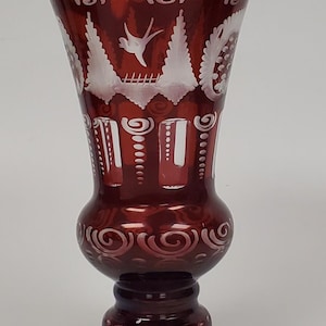 May include: A deep red glass vase with white etched patterns. The vase has a flared top, a rounded middle, and a detailed base. The etched designs include swirling patterns, stylized trees, and a bird in flight, creating a decorative and elegant piece for display.