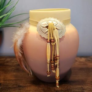May include: A peach-colored ceramic vase with a cream-colored top and a decorative silver-tone starburst buckle. The vase is adorned with leather tassels and feathers, creating a Southwestern or bohemian aesthetic.