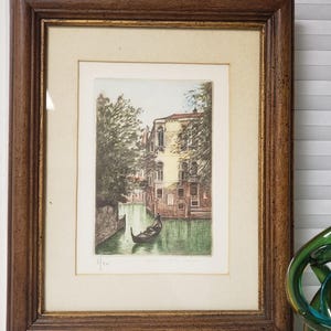May include: A framed etching print depicting a Venetian canal scene with a gondola, a multi-story building, and surrounding trees. The artwork is housed in a dark brown wooden frame with a gold inner trim and a cream-coloured mat board border.